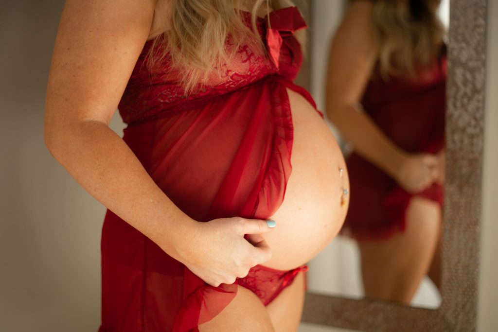 pregnant white woman wearing a red babydoll lingerie set looking down at her baby bump during boudoir maternity photos